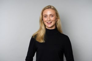 Photograph of Southern Sales Manager Poppy Rules. Poppy is wearing a black turtleneck and has blonde hair which falls past her shoulders. She is smiling towards the camera. She is stood against a plain grey background.