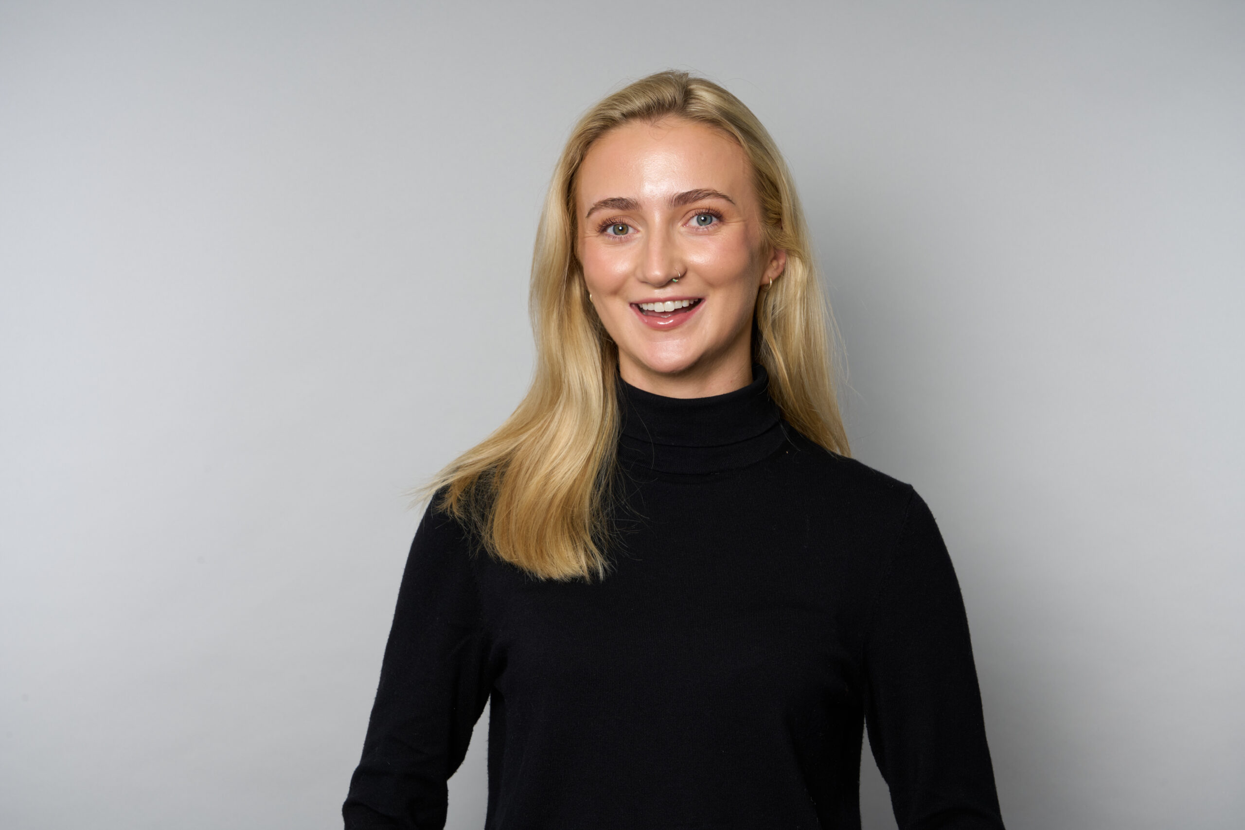 Photograph of Southern Sales Manager Poppy Rules. Poppy is wearing a black turtleneck and has blonde hair which falls past her shoulders. She is smiling towards the camera. She is stood against a plain grey background.