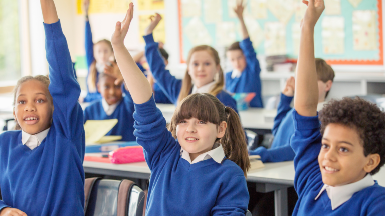 A photograph of school children raising their hands. There are approximately 10 children in the image sat in a well lit classroom. They are wearing blue school jumpers over white shirts.